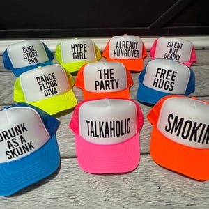 May include: A collection of trucker hats with white fronts and colorful mesh backs. Each hat features a different phrase in bold black letters, including "COOL STORY BRO," "HYPE GIRL," and "SMOKIN."