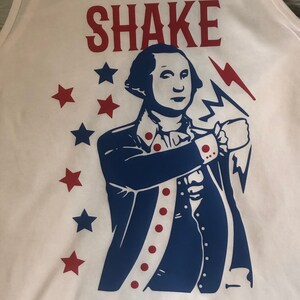 May include: White tank top with a blue and red graphic of a man in a colonial uniform holding a lightning bolt. The text "SHAKE" is printed in red above the graphic. There are red stars around the graphic.