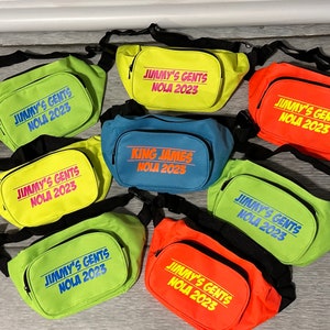 May include: A collection of seven bright colored fanny packs with black straps. The fanny packs have the text "Jimmy's Gents NOLA 2023" printed on them. One fanny pack has the text "King James NOLA 2023" printed on it.