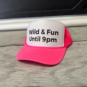 May include: A neon pink trucker hat with a white front panel. The hat features the text "Wild & Fun Until 9pm" in black. The mesh backing is also pink, and the hat is sitting on a wooden surface.