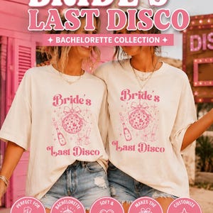 May include: Two off-white t-shirts featuring a pink graphic print of a disco ball with a bow, a champagne bottle, and two glasses. The text reads "Bride's Last Disco". The design includes small star accents. The shirts have short sleeves.