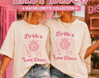 May include: Two off-white t-shirts featuring a pink graphic print of a disco ball with a bow, a champagne bottle, and two glasses. The text reads "Bride's Last Disco". The design includes small star accents. The shirts have short sleeves.