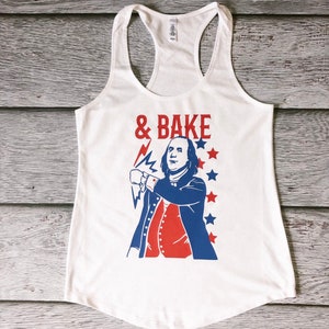 May include: White tank top featuring a red, white and blue graphic of a man with a lightning bolt and the text "& BAKE".