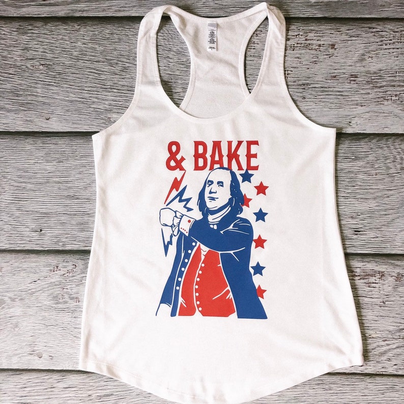 May include: White tank top featuring a red, white and blue graphic of a man with a lightning bolt and the text "& BAKE".