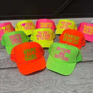May include: A collection of neon trucker hats in various colors, including pink, yellow, and orange. Each hat features bold, humorous text in contrasting colors. The hats have a mesh back and a curved brim, suitable for casual wear.