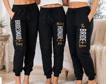 May include: Three pairs of black sweatpants with white and gold text. The left pair reads BRIDESMAID Casey, the middle pair reads Maid of HONOR Jessie, and the right pair reads The BRIDE Lauren. Each features a gold heart and arrow graphic.