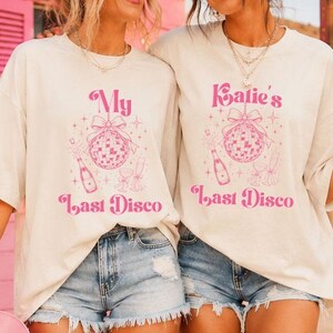 May include: Two off-white short-sleeve t-shirts featuring a pink graphic print. The design includes a disco ball with a bow, a champagne bottle, and glasses, with the text "My Last Disco" on one and "Katie's Last Disco" on the other in a retro font.
