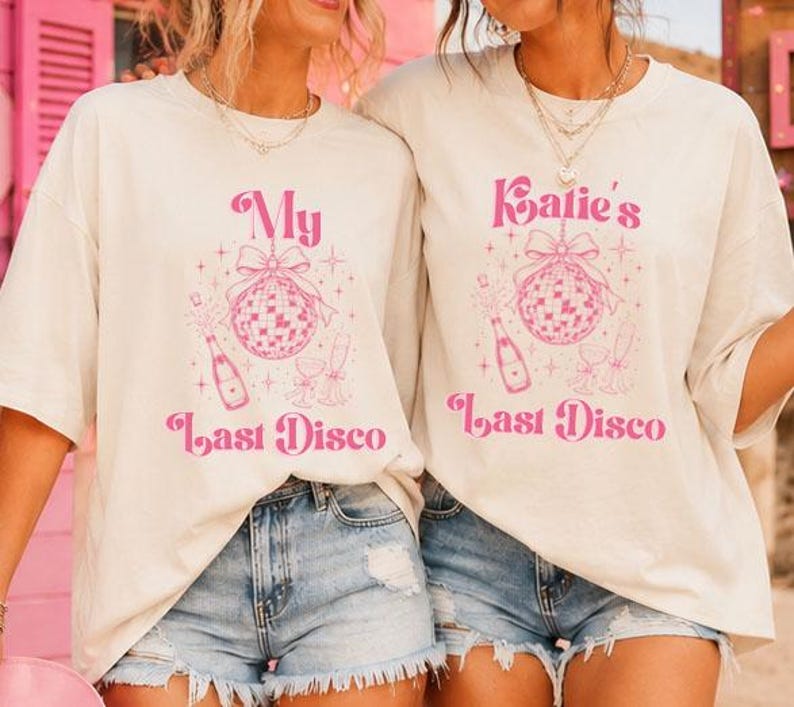 May include: Two off-white short-sleeve t-shirts featuring a pink graphic print. The design includes a disco ball with a bow, a champagne bottle, and glasses, with the text "My Last Disco" on one and "Katie's Last Disco" on the other in a retro font.