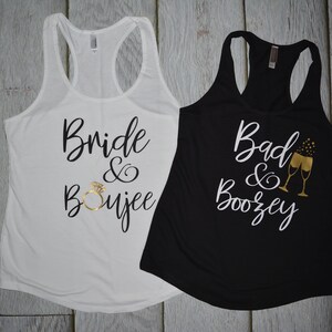 May include: Two tank tops with a bridal theme. The white tank top says "Bride & Boujee" with a gold ring graphic. The black tank top says "Bad & Boozey" with a gold champagne flute graphic.