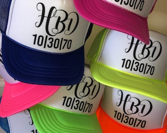 May include: A collection of trucker hats in various colors, including blue, pink, green, and orange. Each hat has a white front panel with the text "HBD 10/30/70" in black script. The hats have mesh backs and colored brims.