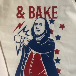 May include: White tank top with a red and blue graphic of a man with a beard and a lightning bolt. The text '& BAKE' is printed in red above the graphic.