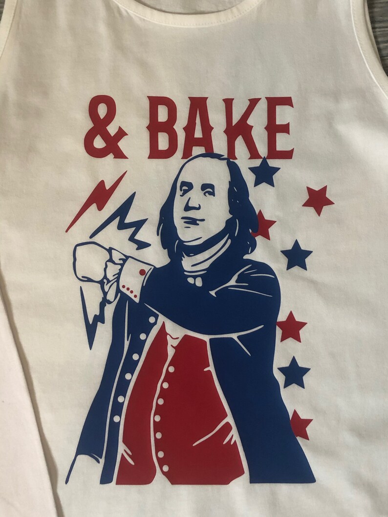 May include: White tank top with a red and blue graphic of a man with a beard and a lightning bolt. The text '& BAKE' is printed in red above the graphic.