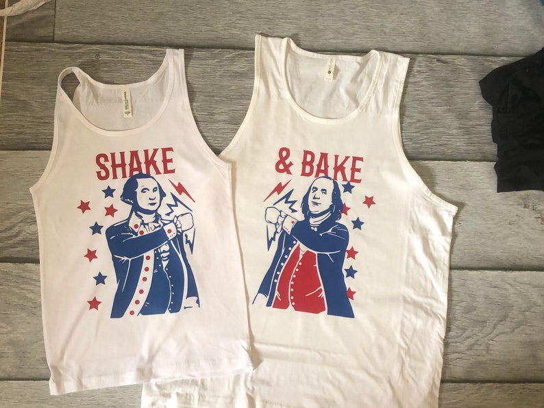 May include: Two white tank tops with a red, white, and blue graphic of a man in a colonial outfit. The text on the tops reads "Shake" and "& Bake".