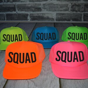 A group of colorful mesh hats with the word "SQUAD" printed on them, arranged on a wooden surface against a brick wall background.