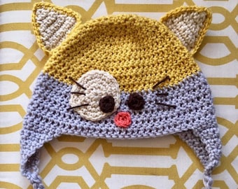 Puede incluir: Un gorro de ganchillo con una parte superior amarilla con dos orejas puntiagudas y una parte inferior gris claro. La cara incluye un parche de ojo color crema, dos ojos circulares marrón oscuro, una nariz pequeña rosa y bigotes bordados.