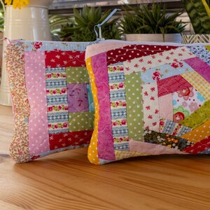 May include: Two colourful, quilted fabric pouches with zip closures. The pouches feature a patchwork design with various floral and geometric patterns in shades of red, pink, blue, green, and yellow. The pouches are rectangular and rest on a wooden surface.