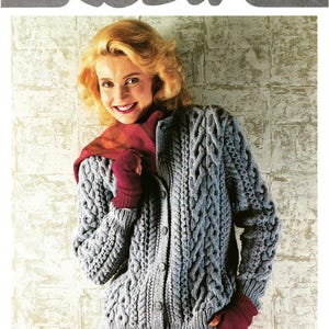 Op de afbeelding: Een vrouw draagt een grijze Aran kabelgebreide cardigan met een knoopsluiting. De cardigan wordt gecombineerd met een rode sjaal en handschoenen. De afbeelding bevat de tekst "Robin" en afmetingen van 81-107 cm.