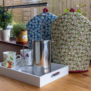 May include: A white serving tray holds a stainless steel French press, two decorative mugs, and a metal mug holder. Two quilted tea cosies with floral patterns and a small potted plant are in the background. The scene is set on a wooden table.