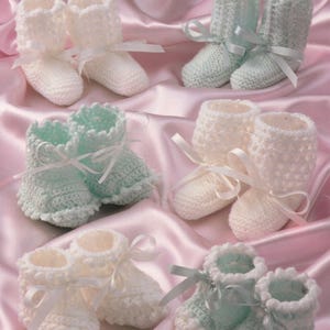 May include: Several pairs of baby booties in various shades of white and light blue are displayed on a pink satin surface. The booties are hand-knitted or crocheted, with delicate ribbon ties. The image also includes the text "8701 BABY 4 PLY Knitting and Crochet".