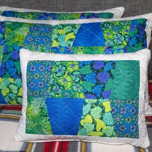 May include: Three rectangular cushions with a patchwork design. The cushions feature a mix of blue and green floral and leaf patterns, framed by a white border. The cushions are arranged on a plaid blanket, creating a cosy and inviting aesthetic.