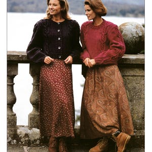 Puede incluir: Anuncio de moda vintage con dos mujeres vestidas con jerséis y faldas largas de punto. Una lleva una rebeca morada oscura y una falda floral, la otra un jersey rojo y una falda con estampado de cachemira. El texto "SIRDAR HIGHLANDER LOPI Chunky" es visible.