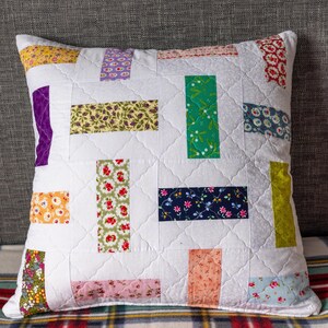 May include: Square white cushion with a patchwork design. It features rectangular fabric patches in various colours and patterns, including floral and geometric prints. The cushion is quilted with a diamond pattern.