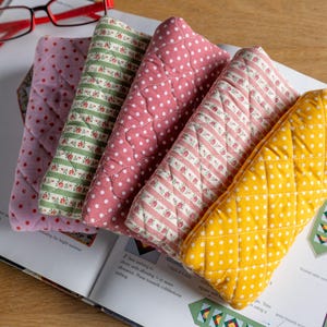 May include: A selection of quilted oven mitts in assorted patterns and colours, including pink polka dots, green floral stripes, and yellow with white dots. The mitts are on an open book, with red-framed glasses visible.