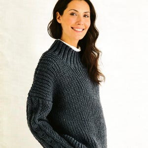 Puede incluir: Un suéter de punto gris oscuro con cuello alto acanalado y mangas de punto trenzado texturizado. El texto Hayfield BONUS 10225 aparece en la parte superior y Bonus with Wool Aran 81 - 137 cm 32 - 54 in está impreso en la parte inferior.