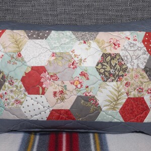 May include: A rectangular cushion with a patchwork design. The centre features a pattern of hexagonal shapes in various floral prints and colours, including red, pink, and green. The cushion has a solid grey border and a quilted texture.