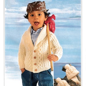 May include: A child wearing a cream-colored cable-knit cardigan with wooden buttons, blue jeans, and a brown bandana. The child carries a wooden stick with a red sack. Two knitted hats are on a piece of driftwood.