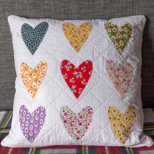 May include: A white cushion featuring nine fabric hearts in various patterns, including floral and polka dots. The cushion has a quilted design and is displayed on a grey surface. The hearts are in different colours.