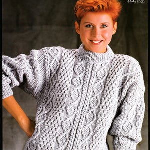 Può includere: Un maglione grigio chiaro in stile Aran con collo alto e intricati motivi a treccia. Il maglione ha un orlo e polsini a coste e le misure sono 84-107 cm. L'immagine include anche il testo "ARAN".