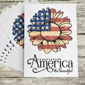 May include: A stack of white cards featuring a sunflower design with the American flag. The sunflower has red and white stripes and blue petals with white stars. The words "America the beautiful" are written in black script.