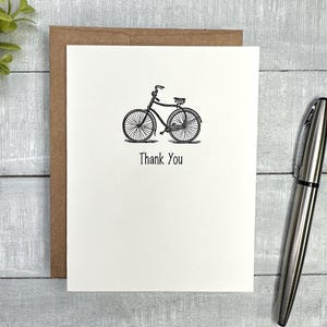 May include: A white card with a black illustration of a bicycle and the words "Thank You" printed below. The card is inside a brown envelope. A silver pen rests on the right side of the card.
