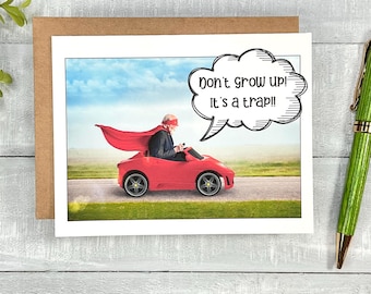 May include: A humorous greeting card featuring a cartoon speech bubble that reads "Don't grow up! It's a trap!" The image shows an older person wearing a red cape and driving a red toy car.