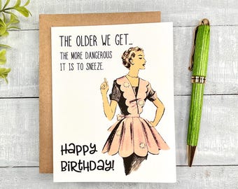 May include: A birthday card with a vintage illustration of a woman in a pink dress, accompanied by the text "THE OLDER WE GET... THE MORE DANGEROUS IT IS TO SNEEZE. Happy BIRTHDAY!" A green pen rests beside the card.