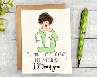 May include: A greeting card featuring a cartoon illustration of a woman holding two pieces of paper. The text on the card reads "You don't have to be crazy to be my friend. I'll train you."