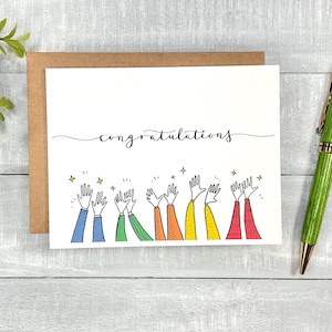 May include: A white card with the word "congratulations" written in black cursive script.  The card features a colorful illustration of seven hands raised in the air, with stars above them.