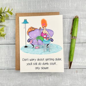 May include: A humorous greeting card with a cartoon illustration of a woman sitting in a purple armchair with a cat. The woman is wearing pink slippers and holding a drink and a snack. The text on the card reads "Don't worry about getting older, you'll still do dumb stuff... only slower."