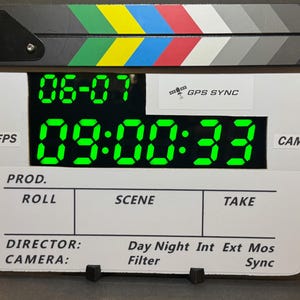 May include: A digital film clapperboard with a black and white body and a multi-colored striped top bar. The screen displays bright green numbers reading 06-07 and 09:00:33. Text on the board includes GPS SYNC, PROD, ROLL, SCENE, TAKE, DIRECTOR, CAMERA, and Filter.