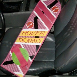 May include: A pink and white hoverboard with the word "HOVER BOARD" in yellow and the "MATTEL" logo. The board has a geometric design with black and white accents and a red strap and green circle.