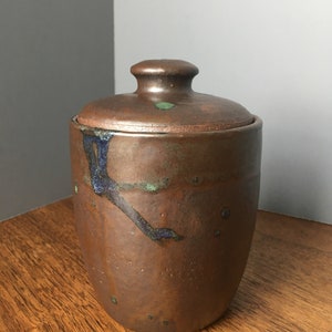 May include: A brown ceramic jar with a lid, featuring a speckled glaze with blue and green accents. The jar has a rounded shape and a smooth, slightly textured surface.