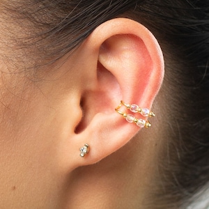 May include: A pair of gold ear cuffs with clear iridescent beads. The cuffs are worn on the upper part of the ear, and there is a small gold stud earring with a clear stone in the lower lobe.