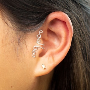 May include: Silver ear cuff with a delicate, swirling design. The cuff is worn on the upper part of the ear, and a small gold stud earring is worn in the lower lobe.