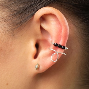 May include: A silver ear cuff with a black bead design. The cuff is worn on the upper part of the ear, and a small gold stud earring is worn in the lower lobe.
