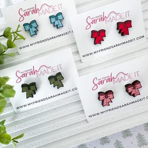 May include: Four pairs of bow-shaped earrings in various colors: light blue, red, olive green, and pink. Each pair is displayed on a white card with the text "My Friend Sarah Made It" and a website address.