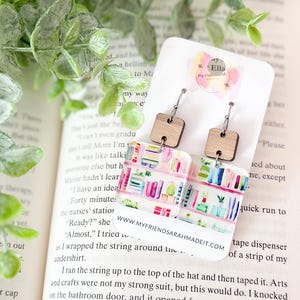 May include: Earrings with a bookshelf design. The earrings feature a wooden square top and a rectangular bottom with a colorful bookshelf print. The earrings are hanging on a white card with the text "Ella" and "www.myfriendsarahmadeit.com".