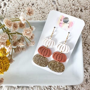 May include: Dangle earrings featuring a pumpkin design. The earrings have white, terracotta, and gold glitter pumpkin shapes. The earrings are attached to gold-colored hoops. The earrings are displayed on a white card.