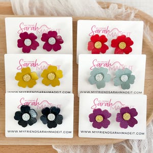 May include: A collection of flower-shaped stud earrings in various colors, including red, yellow, blue-green, black, and purple. Each pair is displayed on a white card with the text "MY FRIEND SARAH MADE IT" and a website address.