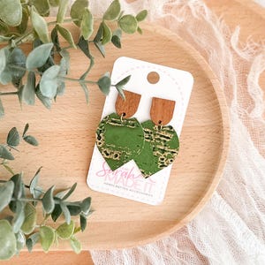 May include: A pair of handcrafted earrings with a green and gold leaf design, topped with a wooden half-circle. The earrings are displayed on a white card with the text "Sarah MADE IT HANDCRAFTED ACCESSORIES". The earrings rest on a wooden tray with greenery.